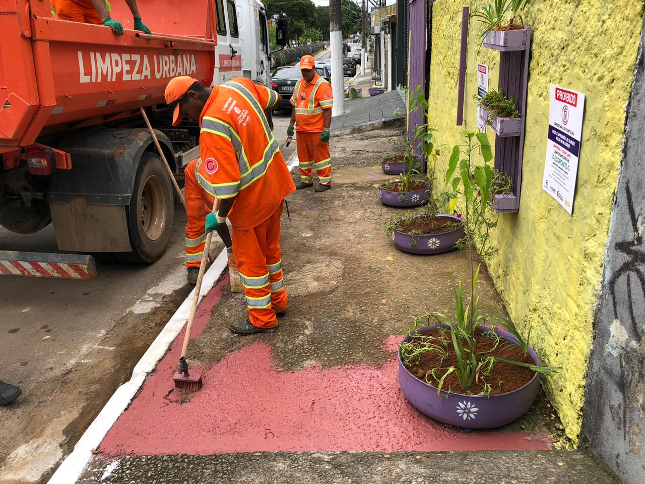 #PraCegoVer visualiza-se funcionários realizando a pintura da calçada do local que está sendo revitalizado. A cor sendo pintada é vinho. Visualiza-se mudas de plantas plantadas no chão.